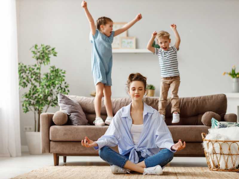 mother-with-closed-eyes-in-lotus-pose-on-the-floor-trying-to-save-inner-harmony-with-children-jumping-on-sofa-and-screaming-in-spacious-room