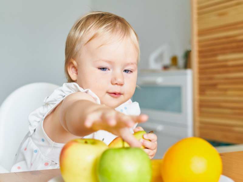 beautiful-baby-on-highchair-reaching-out-for-colorfur-fruits-on-the-table