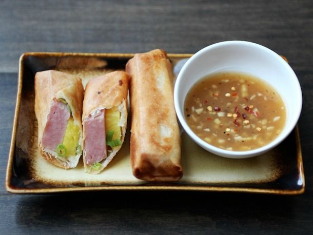 Golden-brown-spring-rolls-filled-with-pineapple-and-ham-on-a-rectangle-plate-served-with-a-small-bowl-of-sweet-and-spicy-sauce.